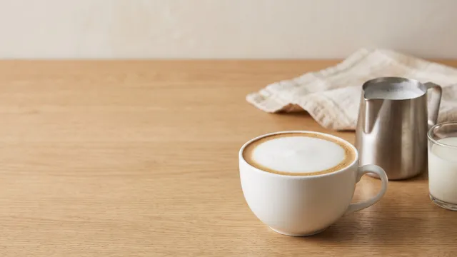 KI-generiertes Symbolbild: weite Tasse mit Milchschaum, Edelstahl-Kännchen und ein Glas Milch auf heller Arbeitsplatte